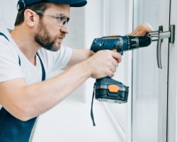 side view of adult repairman in goggles fixing window handle by electric drill